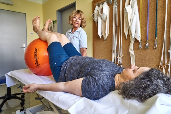 Die Therapeutin erklärt der Rehabilitandin die Übung mit dem Pezziball auf einer Behandlungsliege